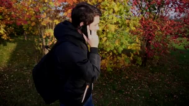 Hombre joven con chaqueta negra caminando en el parque de otoño hablando por teléfono
 - Metraje, vídeo