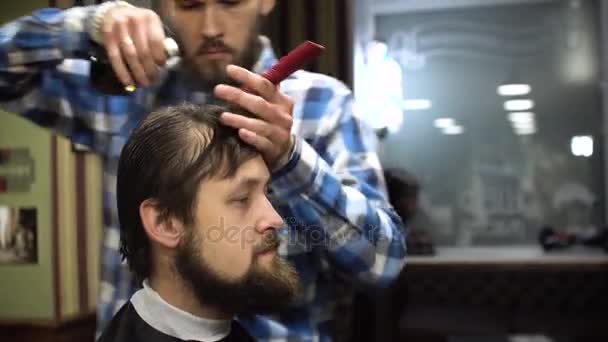 Long hair barber is doing a haircut to young bearded man in barbershop. 4K - Кадры, видео