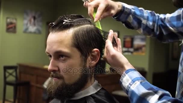 Interior shot of working process in modern barbershop. Close-up portrait of attractive young man getting trendy haircut. Male hairdresser serving client, making haircut using metal scissors and comb - Video, Çekim