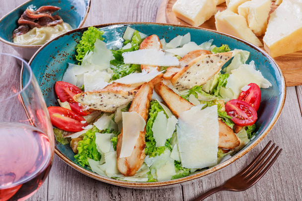 Salad with chicken breast, parmesan cheese, croutons, tomatoes, mixed greens, lettuce and glass of wine on light wooden background. Ingredients on table. Top view - Photo, image