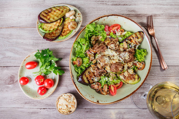 Hot salad with veal, mushrooms, salad leaves, eggplant, zucchini, tomatoes, garnished with grated almonds and Parmesan cheese and glass of wine on wooden background. Healthy food. Top view - Fotoğraf, Görsel