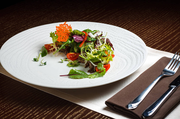 Vegetable salad menu for the restaurant in a large white plate on a white table cloth on a wooden table. Salad on the table with knife and fork. Svezhive vegetables. - Photo, Image