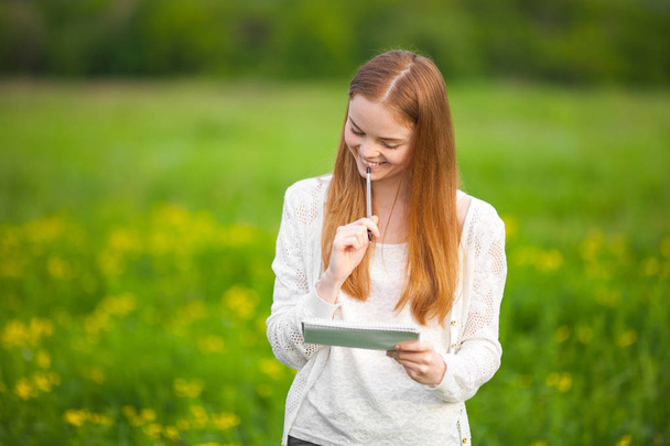 Young freckled meisje landbouwingenieur of bioloog in witte blouse staande in groene veld met notitieboekje en pen tijdens de oogst. - Foto, afbeelding
