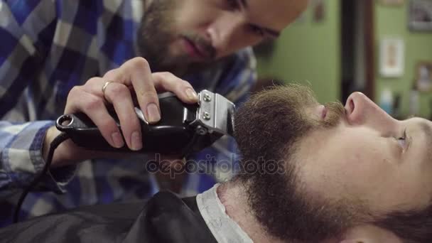 Attractive bearded man getting his beard shaved by modern barber in barber shop - Imágenes, Vídeo