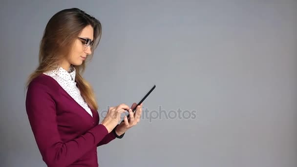 Beautiful young woman in glasses and business casual style using tablet on grey background with a copy space - Footage, Video