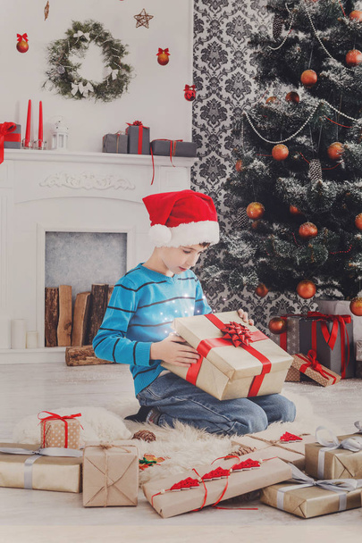 Cute happy boy in santa hat unwrapping christmas presents - Photo, Image