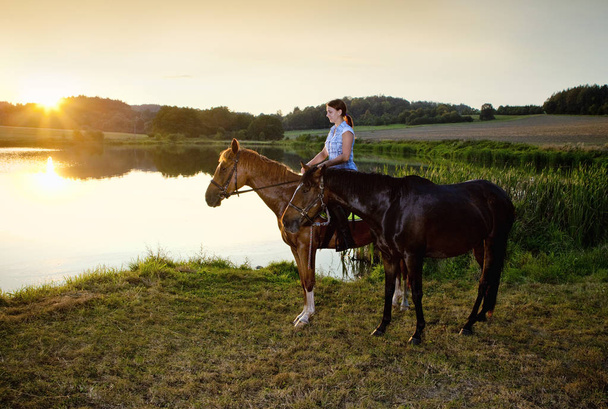 Mulher com dois cavalos por um lago ao pôr do sol
 - Foto, Imagem