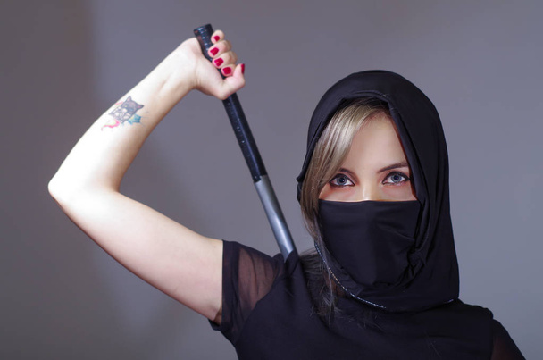 Samurai woman dressed in black with matching veil covering face, holding arm on sword hidden behind back, facing camera, ninja concept - Фото, изображение