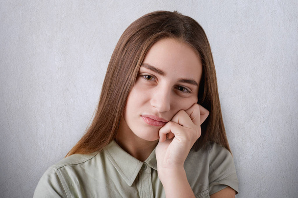 A pretty young girl with brown eyes with full lips and long straight brown hair having a sad look and keeping her hand on her cheek. A portrait of an attractive grieved girl on the white background  - Foto, Imagen
