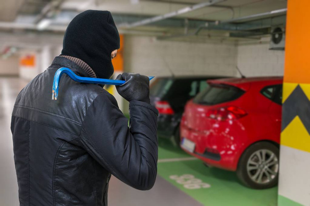 Thief in balaclava holds crowbar in hand and is going to steal a car from parking lot. - Photo, Image