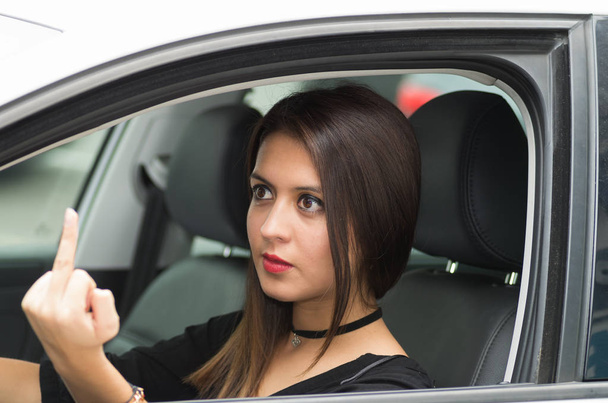 Closeup νεαρή γυναίκα που κάθεται στο αυτοκίνητο δίνει το δάχτυλο θυμωμένα, όπως φαίνεται από έξω οδηγών παράθυρο, γυναίκα οδηγός έννοια - Φωτογραφία, εικόνα