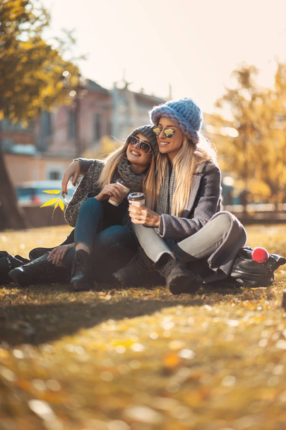 Two beautiful happy women sitting in the park on grass drinking coffee and laughing. Concept friendship, togetherness, happiness. - Фото, зображення