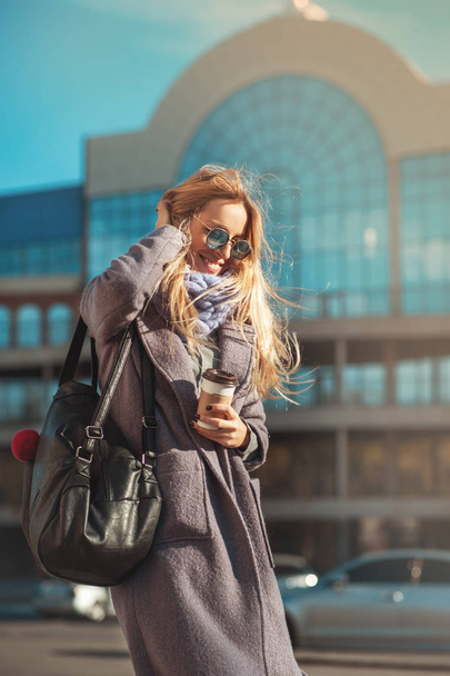 Отличная погода сегодня для прогулки. Красивая молодая блондинка держит чашку кофе и гуляет на улице с улыбкой на лице
 - Фото, изображение