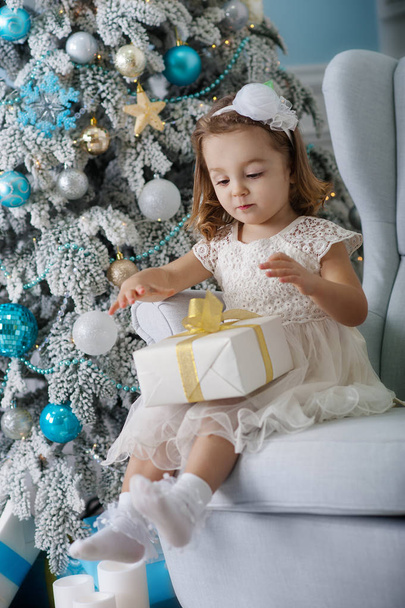 cute little girl in bklom dress sitting in a chair and opens  box with  present for background Christmas tree  blue ornaments - Foto, Imagem