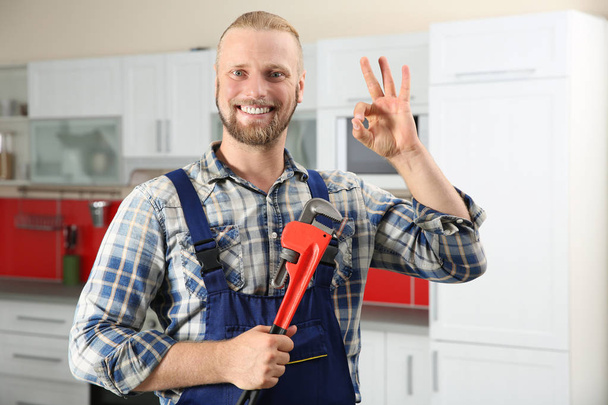 Portrait of handsome plumber with pipe wrench showing OK sign in kitchen - Foto, Bild