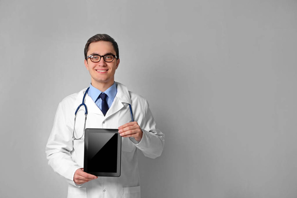 Young male doctor with tablet on grey wall background - Fotoğraf, Görsel