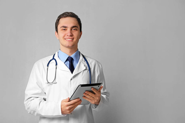 Young male doctor with tablet on grey wall background - Zdjęcie, obraz