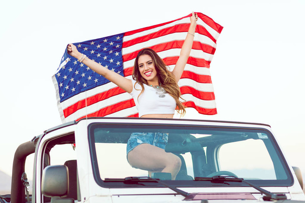 glückliches junges Mädchen steht in Cabrio-Auto und schwenkt amerikanische Flagge. - Foto, Bild