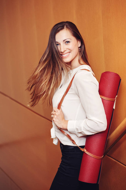 Portrait of beautiful smiling young white Caucasian business woman with blue eyes in white blouse shirt and black pants holding, carrying yoga mat in office, standing on staircase, lifestyle concept - Photo, Image