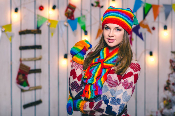 Young beautiful european girl in colored knitted hat and gloves. Woman smiling, standing on the background of Christmas decorations. - Fotó, kép