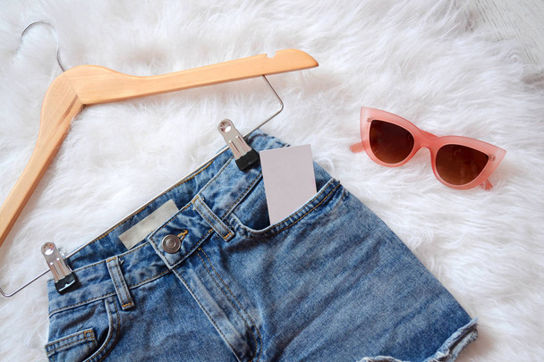 kurze Jeans-Shorts auf Trempel mit leerer Visitenkarte in der Tasche, auf weißem Pelz und rosa Brille. Blick von oben - Foto, Bild