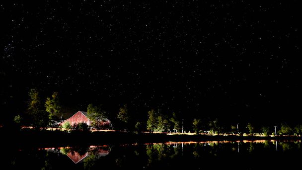Cena noturna de um lago no Canadá com um celeiro vermelho e luzes de carros que passam
 - Foto, Imagem