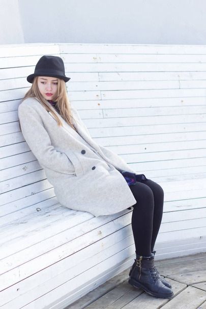 lonely sad beautiful girl in a black coat and hat, sitting on a white bench cold winter sunny day - Фото, изображение