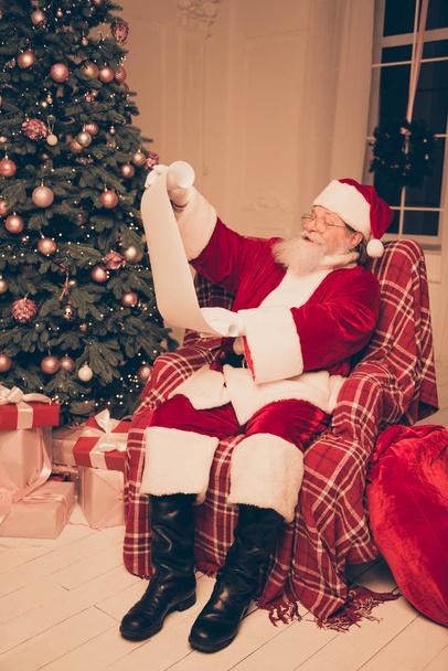 Santa Claus wearing red costume and black shoes holding list of  - Foto, Imagen