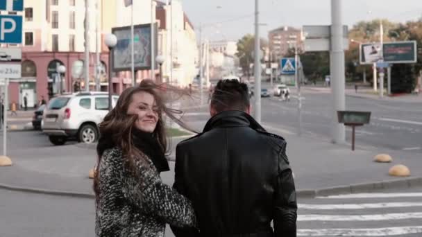 Back view of happy lovers walking in the street. Beautiful girl looks back into camera smiling. Slow mo, steadicam shot - Imágenes, Vídeo