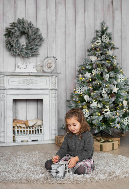 Pretty little girl sits on a floor and plays with wooden toy near a fireplace in Christmas at home - Φωτογραφία, εικόνα