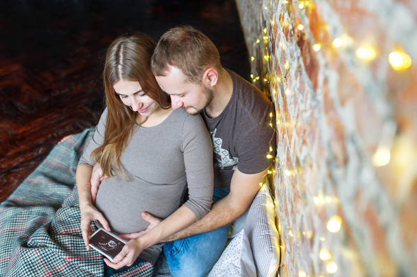 Vista superior de una joven pareja sentada en el suelo, esperando un bebé. Consideran la foto ultrasonografía
. - Foto, Imagen