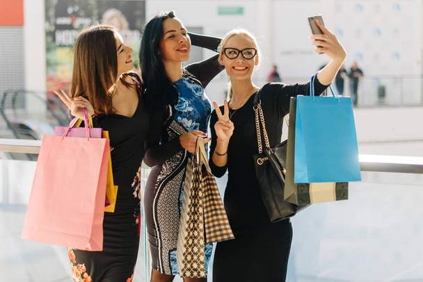 Attractive pretty women taking selfie after shopping. Girls are very happy, smiling. They posing for photos. Have many colorful shopping bags in hands - Photo, image