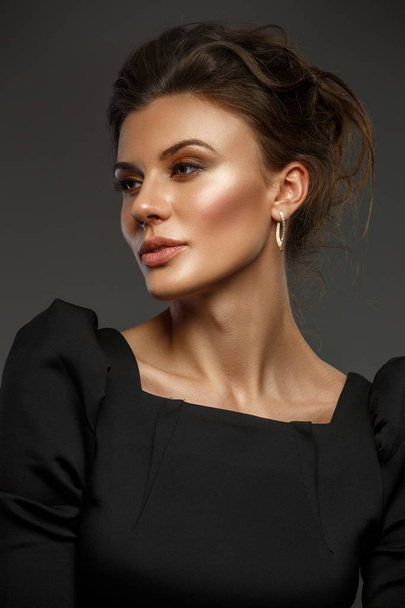 Portrait of a beautiful, self-sufficient and confident young woman with collected hair in the elegant black dress with makeup, dark background, studio photo - Foto, imagen