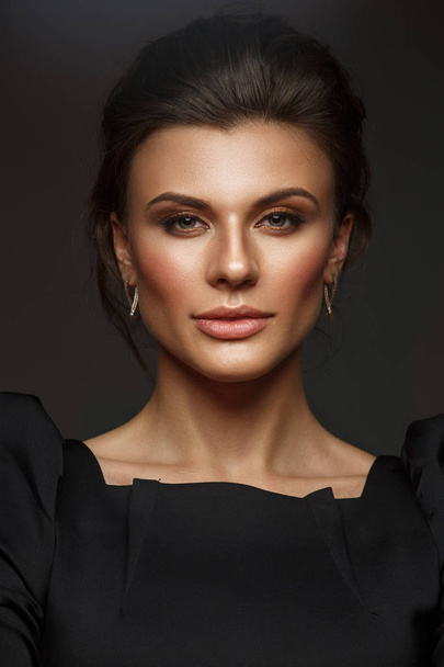Portrait of a beautiful, self-sufficient and confident young woman with collected hair in the elegant black dress with makeup, dark background, studio photo - Valokuva, kuva