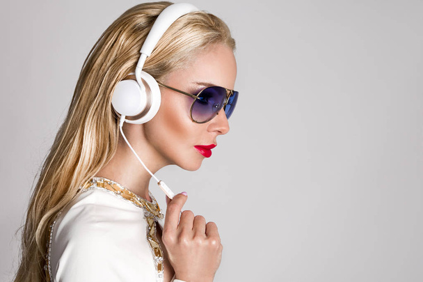 Beautiful sexy blonde woman with long hair and perfect body in an elegant white suit sitting with headphones on and listen to music  - Foto, immagini