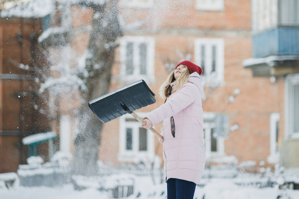 Νεαρό κορίτσι καθαρίζει χιόνι κοντά στο σπίτι, κρατώντας ένα φτυάρι και κουπί περνούν το χρόνο - Φωτογραφία, εικόνα