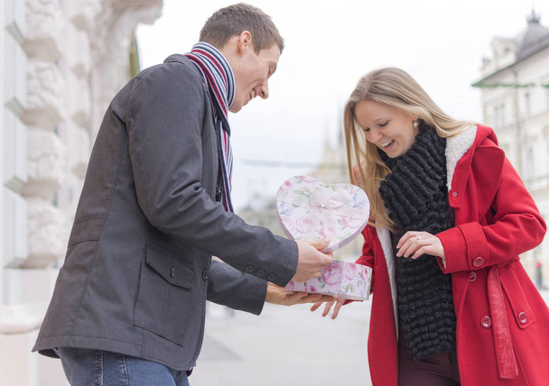 schöner junger Mann, der seiner Freundin eine Geschenkbox schenkt, während er auf der Straße steht. Liebe und Valentinstag-Konzept. - Foto, Bild