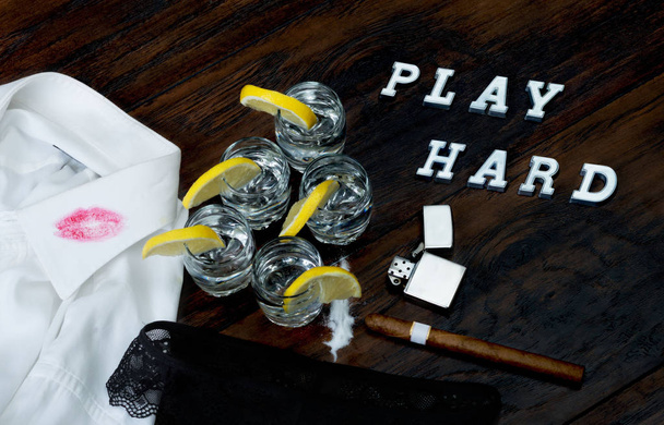 Jogar duro escrito na mesa de madeira com tequilas, charuto cubano, isqueiro, camisa e roupa interior mulher
.  - Foto, Imagem