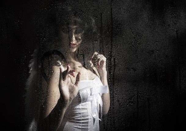 Beautiful bride wearing white dress with professional make-up and hairstyle, posing behind transparent glass covered by water drops. wings behind - Valokuva, kuva