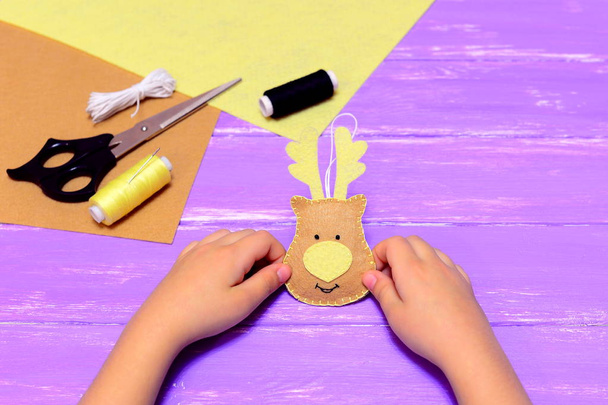 Little child holds a felt Christmas deer decoration in hands. Yellow and brown felt pieces, thread, needle, scissors on lilac wooden background. Christmas sewing handicrafts concept  - Photo, Image