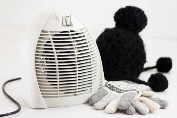 Felmelegedés szoba, télen, a kesztyű és a gyapjú sapka fehér padlón elszigetelt elektromos ventilátor melegítő - Fotó, kép
