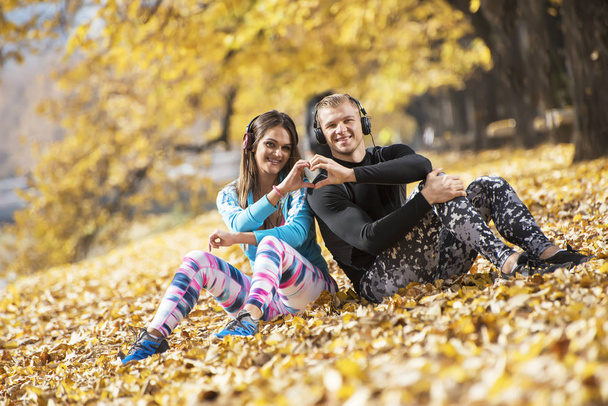 Belo jovem casal descansando e ouvindo música após o treinamento bem sucedido no parque. Coração em forma de mão. Ambiente de Outono
. - Foto, Imagem