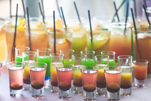 Beautiful row line of different colored alcohol cocktails on a party, martini, vodka,and others on decorated catering bouquet table on open air event, picture with beautiful bokeh - Фото, изображение