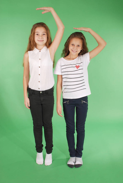 Kaksi koulutyttöä pitää toisiaan kädestä pään päällä ja mittaa hänen kasvuaan.
 - Valokuva, kuva