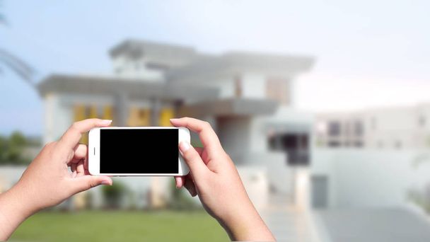 Hand holding white mobile smart phone over blurred house backgro - 写真・画像