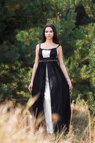 Грациозная очаровательная брюнетка, позирующая в черном платье в хвойном парке
 - Фото, изображение