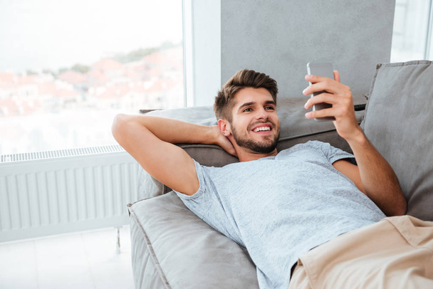 Happy young man lies on bed and looking at phone - 写真・画像