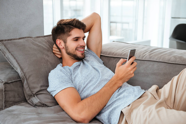 Happy young man lies on sofa and chatting by phone - Фото, изображение