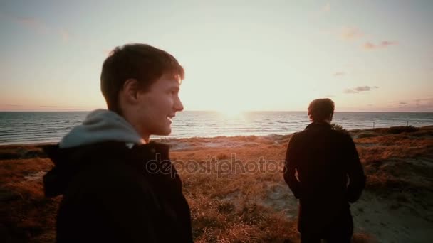 Dos jóvenes están de pie en las dunas de arena en la playa disfrutando de la vista de una hermosa puesta de sol
 - Imágenes, Vídeo