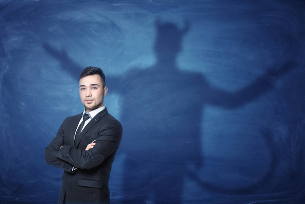 Businessman standing with hands across and his shadow on blue blackboard behind him tail like devil - Φωτογραφία, εικόνα